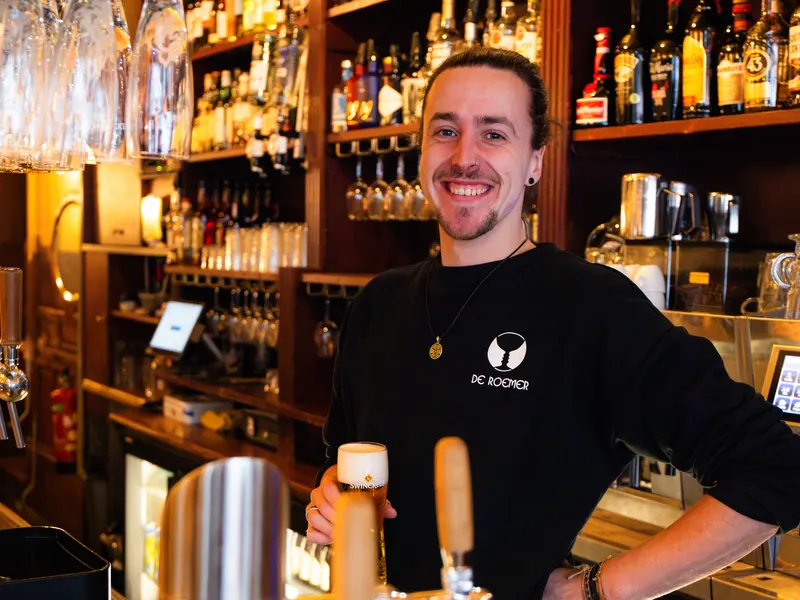 Een medewerker tapt lachend een glas bier achter de bar.