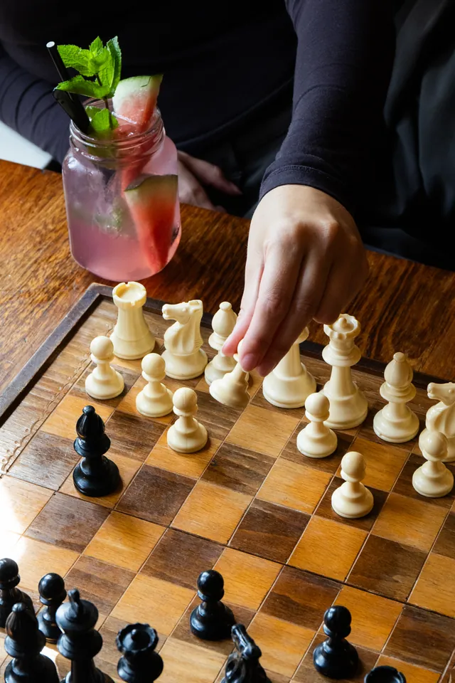 Hand verzet een schaakstuk op een houten schaakbord, met daarnaast een rode cocktail met munt.