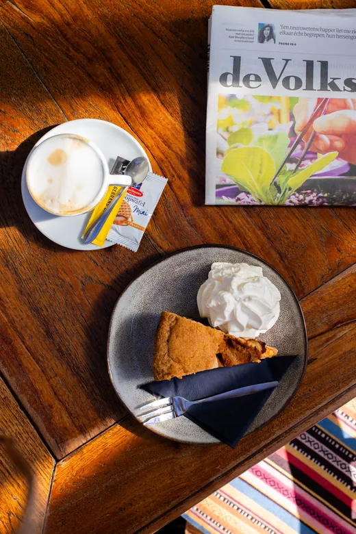 Cappuccino met een punt appeltaart met slagroom naast een opengeslagen krant.