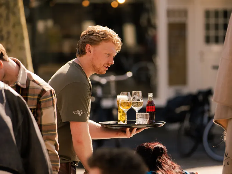 Een medewerker loopt met een dienblad vol bier en frisdrank langs het terras.